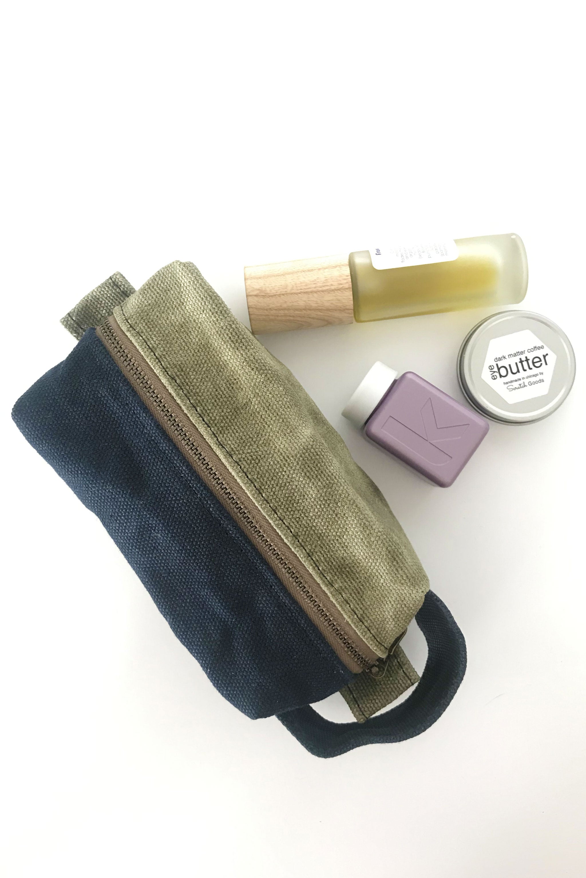 small waxed canvas toiletry bag with handle and brass zipper. The bag is closed and sits next to three small bottles and containers. The bag is half sage green and half dark navy blue. 