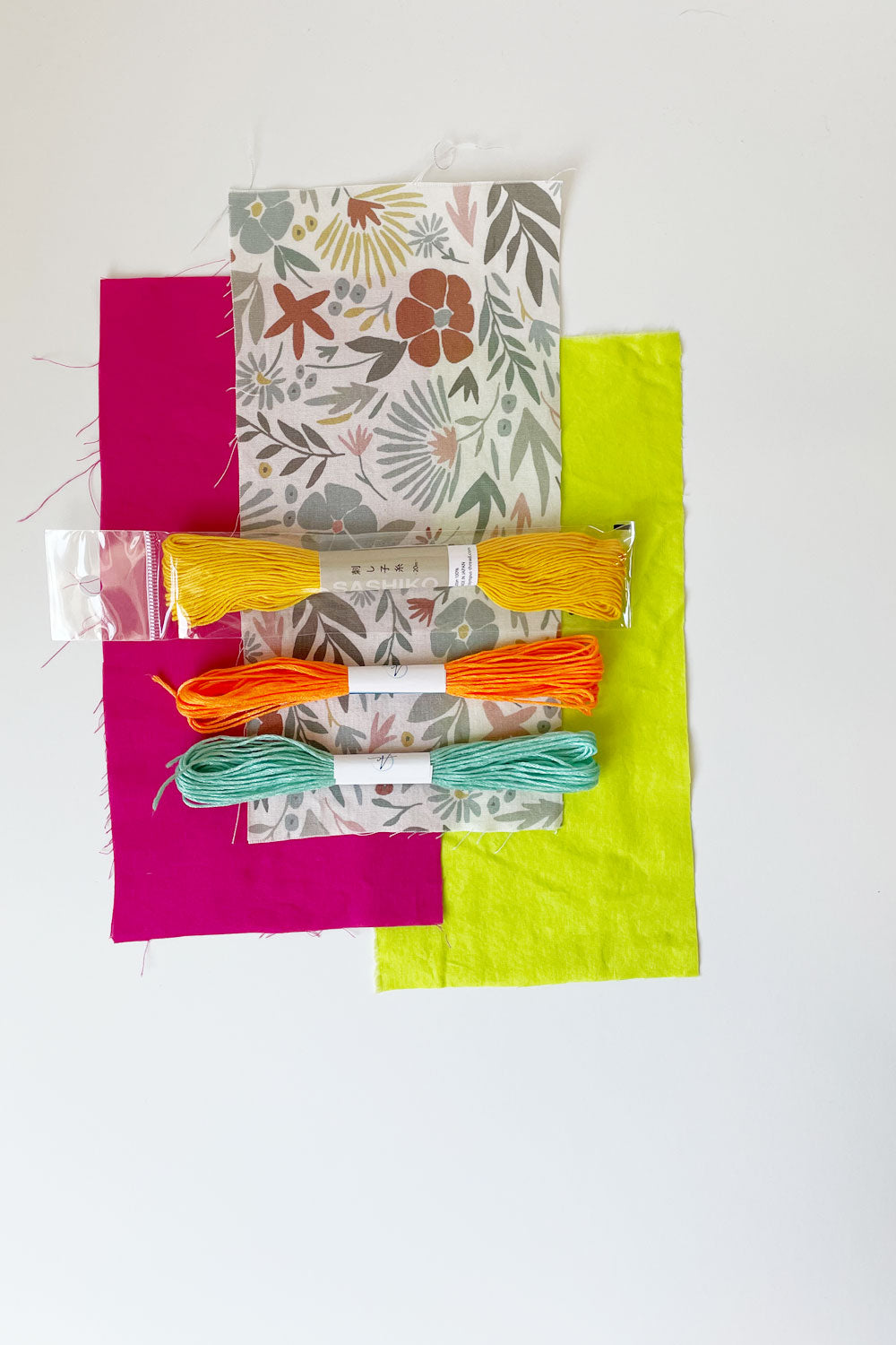 3 fabric swatches with sashiko thread and 2 embroidery floss laying on top. Floral print colors are rust, yellow, and steel blue on white background. Bright fuchsia and neon light green fabrics. Sashiko thread in yellow with embroidery threads in orange and sea foam green.
