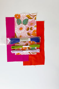 3 fabric swatches with sashiko thread and 2 embroidery floss laying on top. Floral print colors are light pink, orange, and green on light pink background. Bright fuchsia and orange fabrics. Sashiko thread in ultramarine blue with embroidery threads in lime green and light purple.