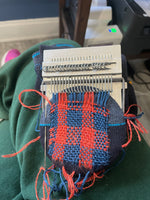 Load image into Gallery viewer, A close-up of a hand-woven darn sample in progress, showing a red and blue basketweave pattern, held in place by a metal knitting or weaving frame. The sample is positioned on a person's lap wearing green pants, with an office background visible.

