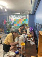 Load image into Gallery viewer, Three people sitting at tables with sewing machines and a colorful wall window display in the background.

