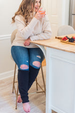 Load image into Gallery viewer, woman in beige sweater and jeans with black leg warmers cover her legs below the knees. She is smiling while holding a glass of rose wine, sitting at a bar stool with a plate of fruit next to her. 

