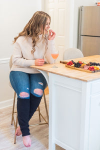 woman in beige sweater and jeans with black leg warmers cover her legs below the knees. She is taking a bite of fruit while a glass of rose wine sits on the counter. She is sitting at a bar stool with a plate of fruit next to her. 