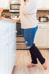woman stands in kitchen with white cabinets and tan floors. She is wearing a beige knit sweater, jeans, and black leg warmers cover her leg below the knee. A glass of rose wine and a fruit plate sit on the counter. 