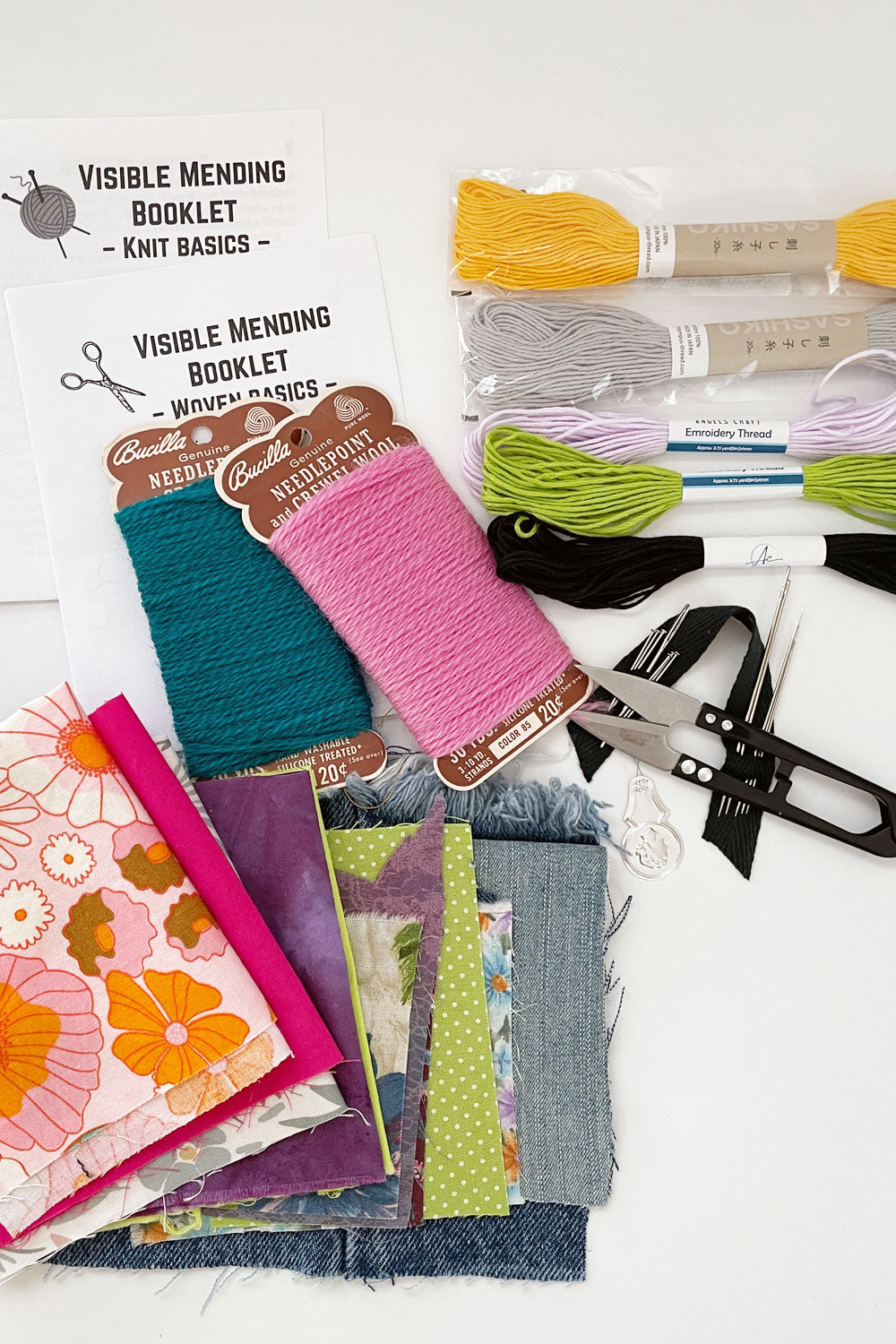 Assortment of mending supplies: Visible mending booklets for knit and woven fabrics made from paper. 2 sashiko threads, 2 embroidery threads, 2 needlepoint crewel wool cards, thread snips, needles and pins on cotton twill tape, and small assortment of cotton and denim fabric scraps. 