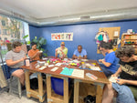 Load image into Gallery viewer, Group of people sitting and hand sewing around a table in a casual setting with sewing supplies, tool, and materials items on it.

