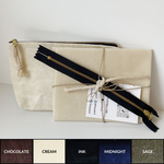 Load image into Gallery viewer, Pre-cut kit with waxed fabric, zipper, zipper pull, and QR code tied with twine leans against a sewn cream colored waxed zippered pouch. Available colors of fabric shown with text: chocolate (brown), cream (off-white), ink (black), midnight (navy blue), sage (green. 

