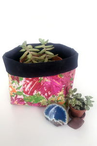 Hot pink floral with bright green, orange, yellow, and light pink abstract design on a fabric basket. Solid black lining. The lining is folded down an inch. A small succulent pot sits inside the basket. There is a small succulent in a brown plastic container next to a blue geode and smooth brown stone. 