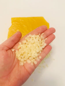A hand holds a small pile of beeswax pellets. A block of beeswax is in the background on a white background. 