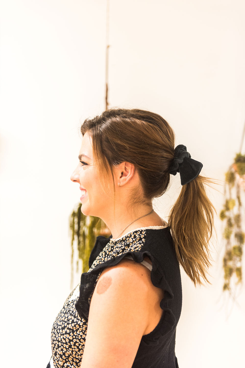 Side view of a woman with her hair tied in a mid-ponytail with a black velvet bow hair scrunchie. She is smiling. 