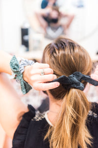 Back view of a woman tightening her ponytail. She wears a black velvet bow hair scrunchie in her hair. Two more hair scrunchies are on her left wrist. 