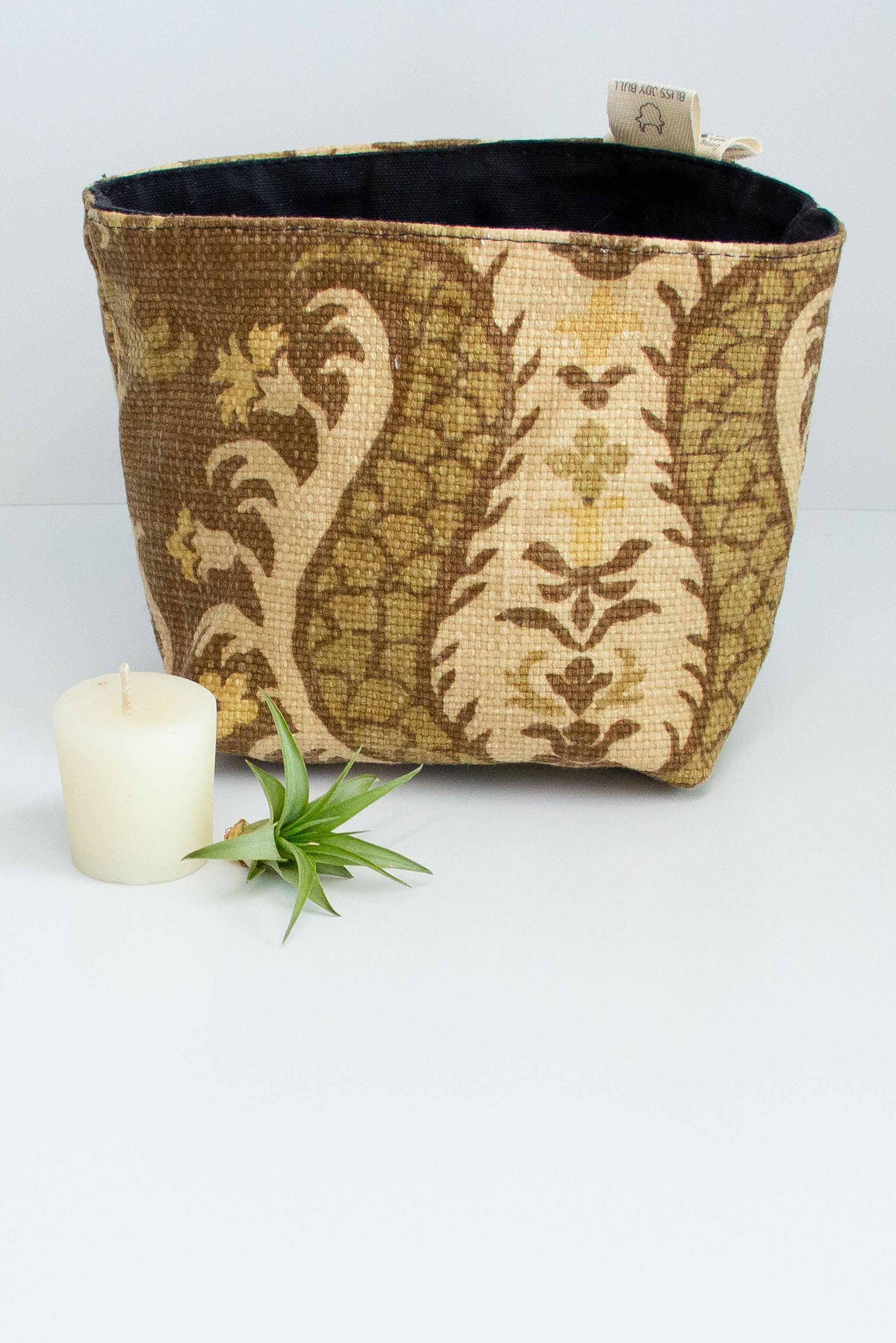 A fabric basket in light yellow and dark olive fleur de lis pattern with a black lining. A small votive candle and air plant sit next to it on a white background. 