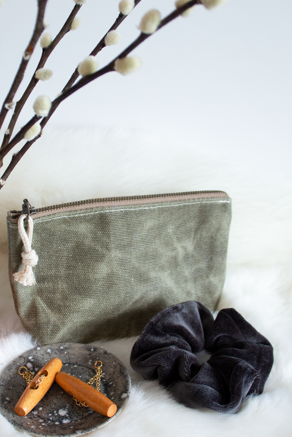 Zippered pouch on faux fur with black velvet hair scrunchie, wood and brass earrings on a ceramic tray, and pussy willows. 
