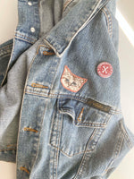 Load image into Gallery viewer, Sad cat head applique on denim jacket with pin that reads community glue workshop. 
