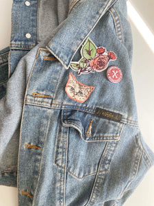 Sad cat head applique and rose bush patch on denim jacket above the left pocket with pin that reads community glue workshop. 