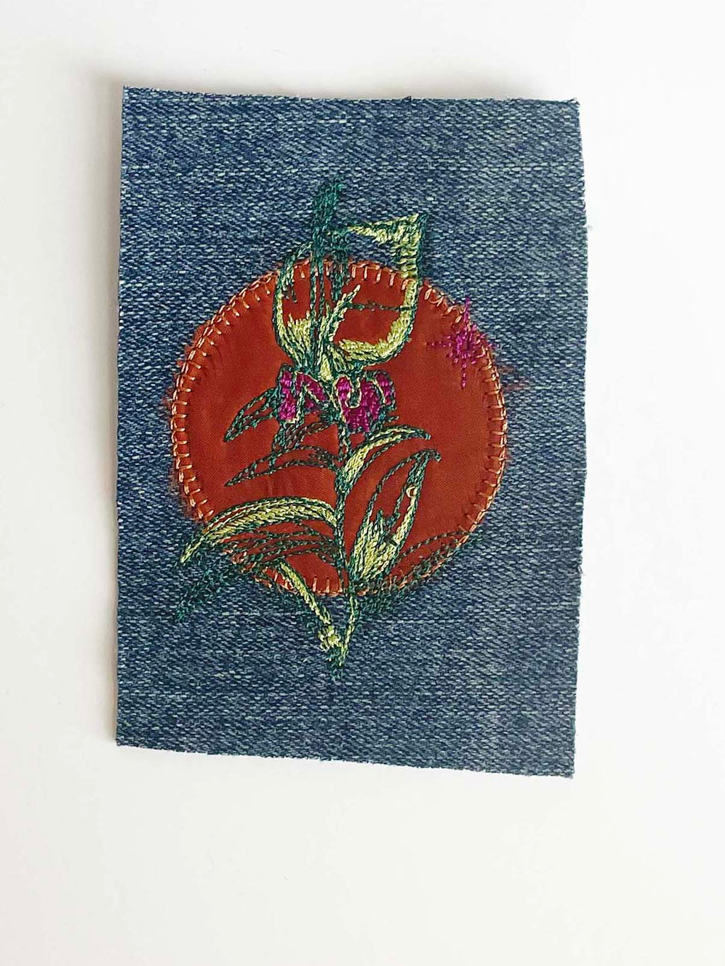Machine embroidered bouquet of flowers in shades of magenta with neon green and dark green stem. Copper circle fabric is embroidered behind the flowers on a rectangle piece of denim. 