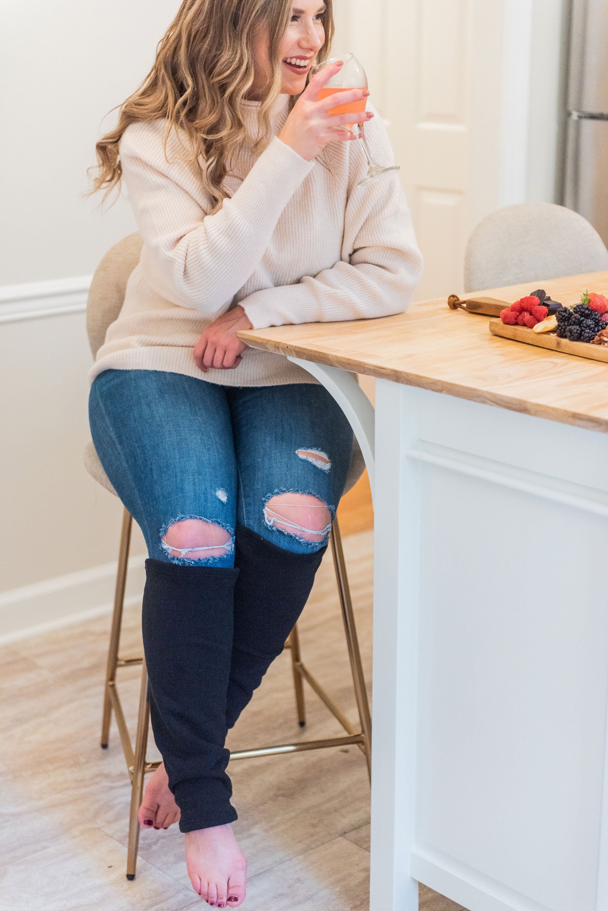 woman in beige sweater and jeans with black leg warmers cover her legs below the knees. She is smiling while holding a glass of rose wine, sitting at a bar stool with a plate of fruit next to her. 