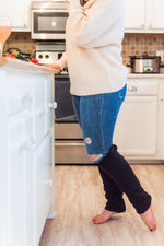 Load image into Gallery viewer, woman stands in kitchen with white cabinets and tan floors. She is wearing a beige knit sweater, jeans, and black leg warmers cover her leg below the knee. A glass of rose wine and a fruit plate sit on the counter. 
