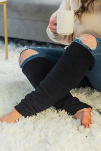 woman sits on a white plush fuzzy carpet while holding a white mug of tea. She is wearing a beige knit sweater, jeans, and black leg warmers that cover her legs below the knee.