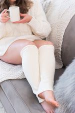 Load image into Gallery viewer, woman in cream chunky knit sweater sits on grey couch with cream knit blanket, holding a white mug of tea and wearing cream leg warmers below the knee
