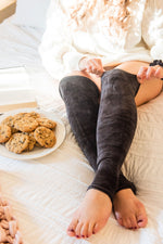 Load image into Gallery viewer, woman sits on cream comforter with her ankles crossed. She wears a chunky knit white sweater and black velvet leg warmers. She is adjusting one of the leg warmers. A plate of chocolate chip cookies and open book sit next to her on the bed. 
