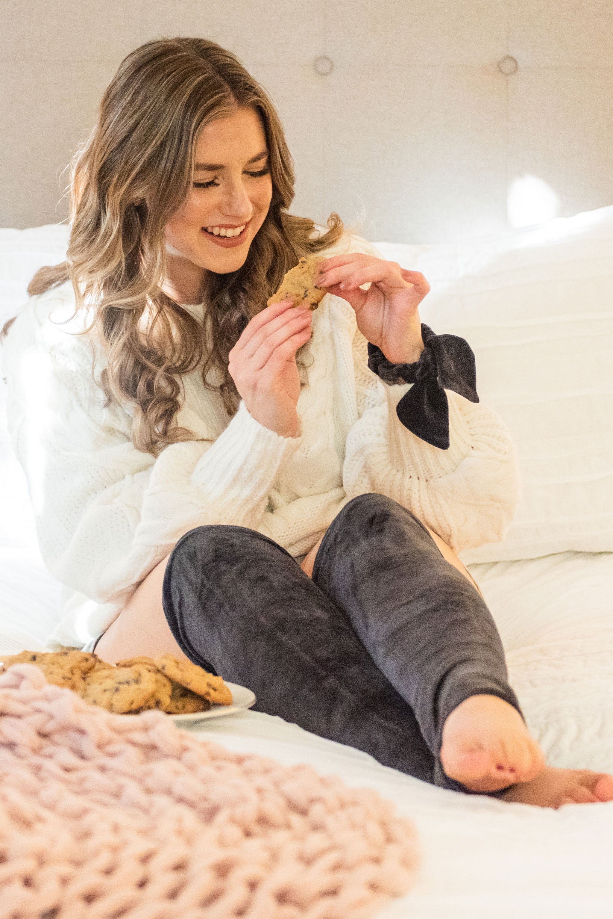 A smiling woman sits on a bed with white bedding. She is wearing a white chunky knit sweater and black velvet leg warmers. She is wearing a bow hair scrunchie on her left wrist. She is holding a partially eaten cookie about to take another bite. A plate of cookies and pink chunky knit blanket are on the bed next to her..