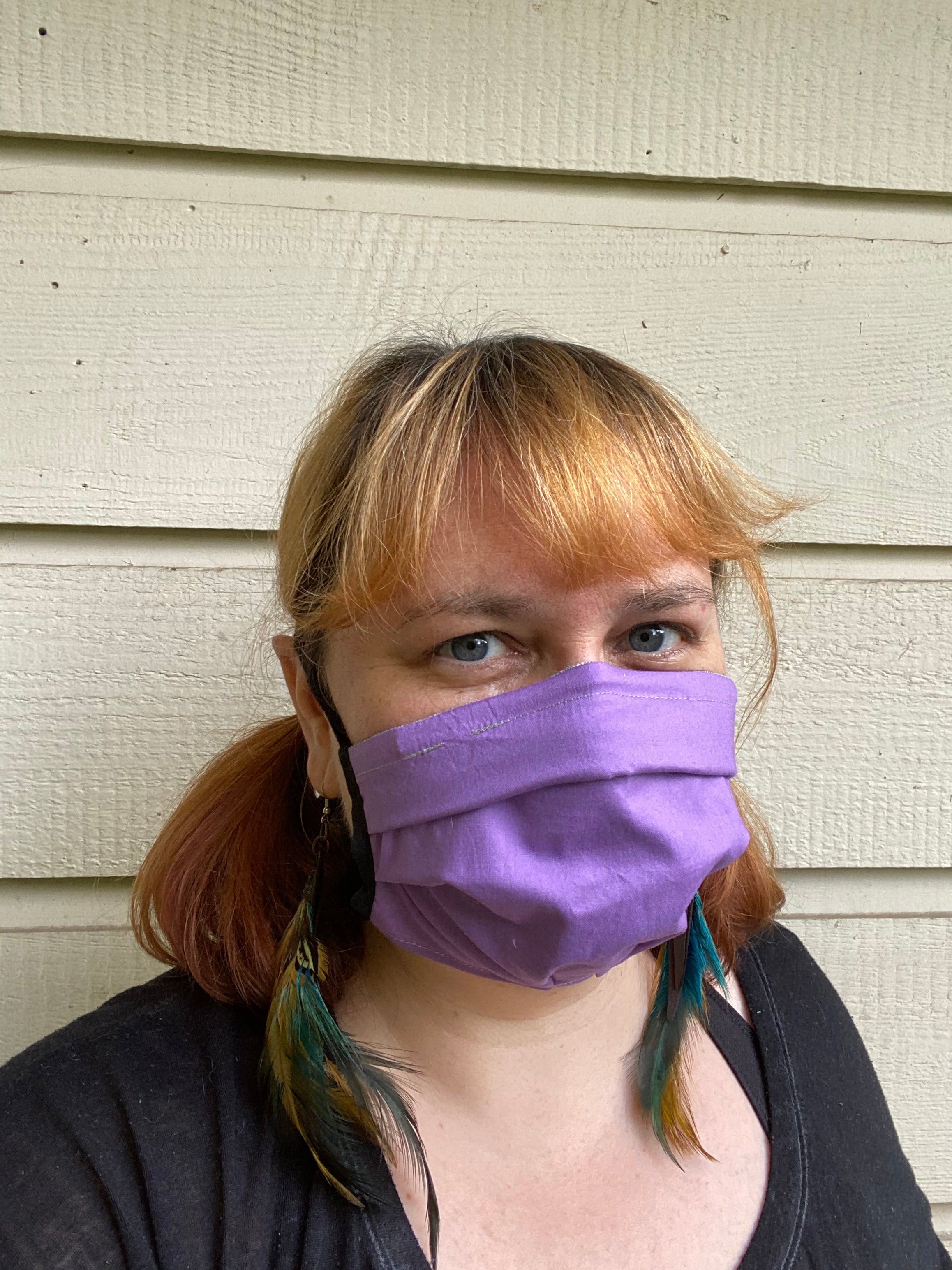 Woman wears a purple fabric face covering, feather earrings, and black t-shirt. She is smizing behind her mask. This is a portrait view of her. The background is a beige painted wood siding. 