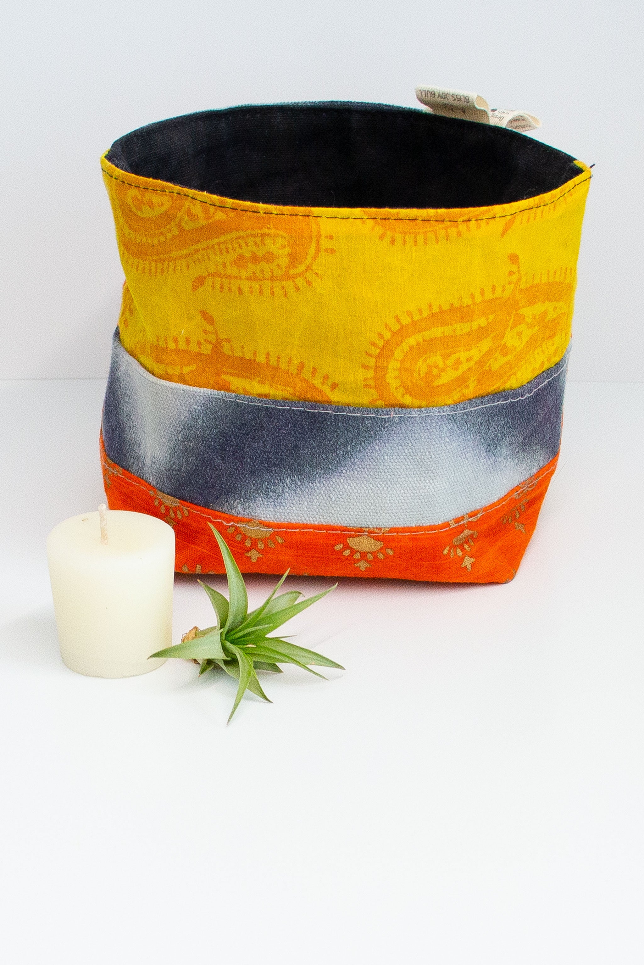 Patchwork fabric basket in yellow with darker yellow paisley pattern, with with dip dyed blue pattern, and orange with gold pattern. Solid black lining. A small votive candle and air plant sit next to it on a white background. 