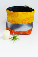 Load image into Gallery viewer, Patchwork fabric basket in yellow with darker yellow paisley pattern, with with dip dyed blue pattern, and orange with gold pattern. Solid black lining. A small votive candle and air plant sit next to it on a white background. 
