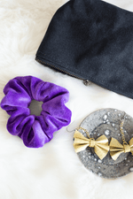 Load image into Gallery viewer, Purple velvet scrunchie, black zippered pouch, and gold bow earrings on grey ceramic holder, all on faux fur background. 
