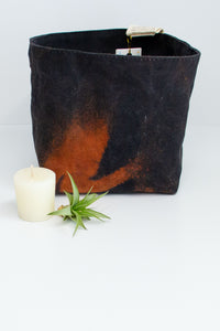 A black fabric basket is discharged dyed, which creates an abstract pattern in a rust color. A small votive candle and air plant sit next to it on a white background. 