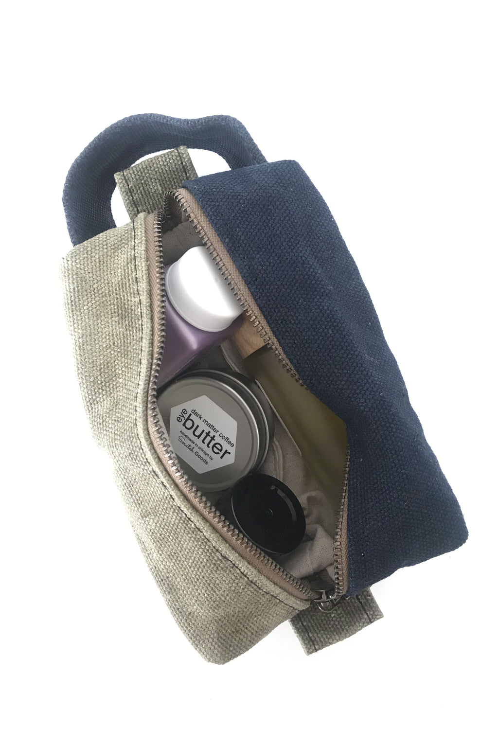 small waxed canvas toiletry bag with handle and brass zipper. The bag is open and contains small toiletries inside. The bag is half sage green and half dark navy blue. 