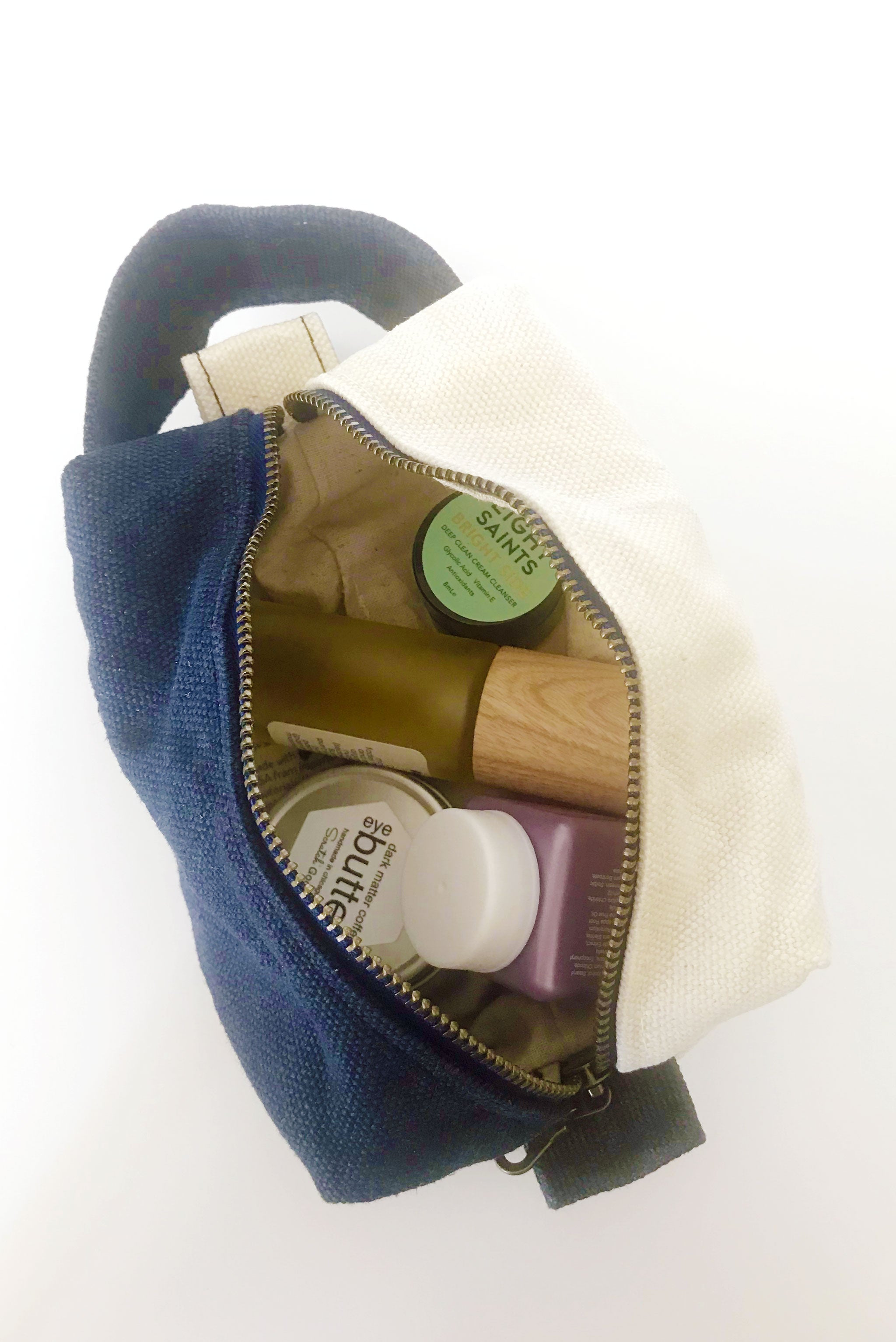 small waxed canvas toiletry bag with handle and brass zipper. The bag is open and contains small toiletries inside. The bag is half cream and half dark navy blue. 