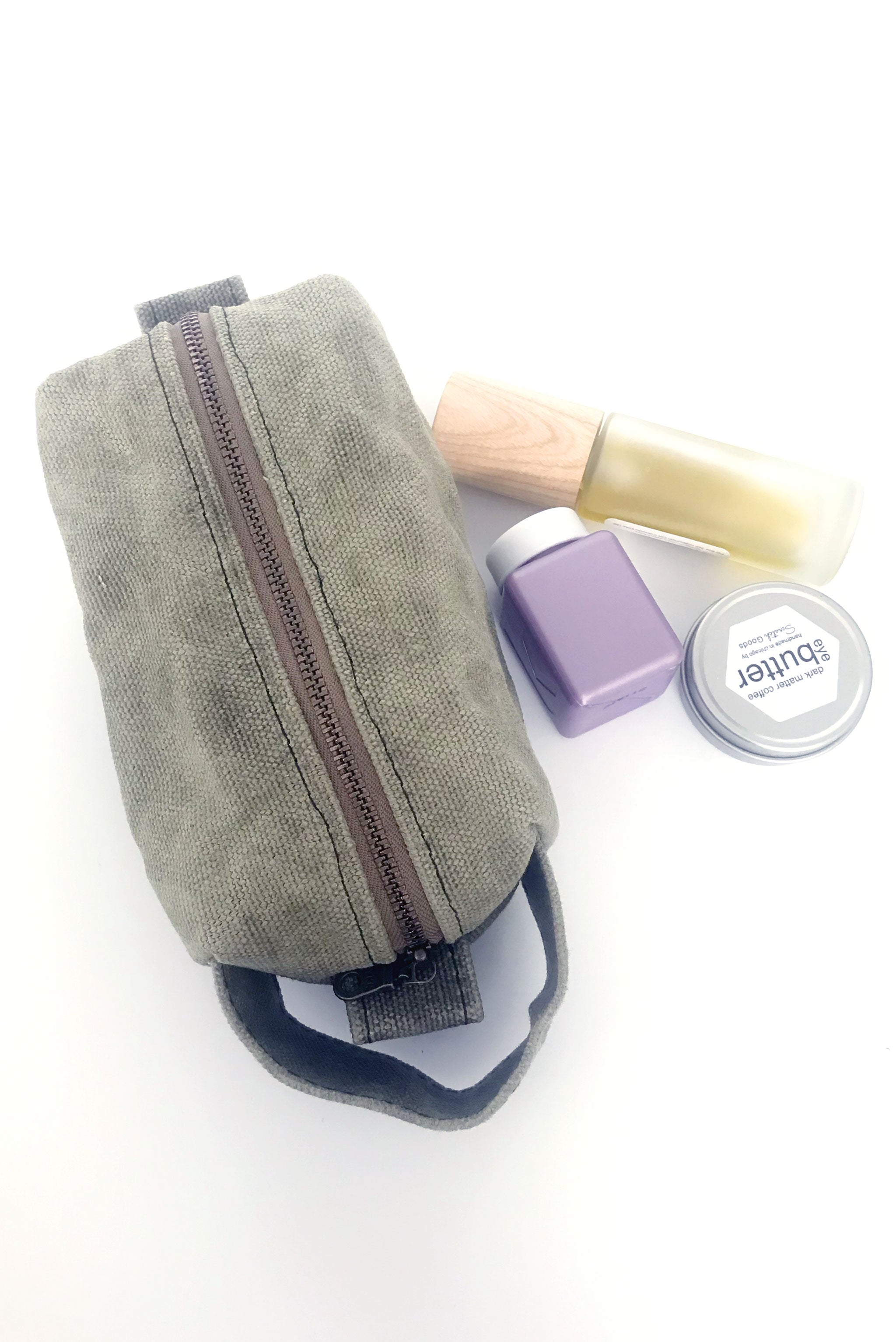 small waxed canvas toiletry bag with handle and brass zipper. The bag is closed and sits next to three small bottles and containers. The bag is sage green. 