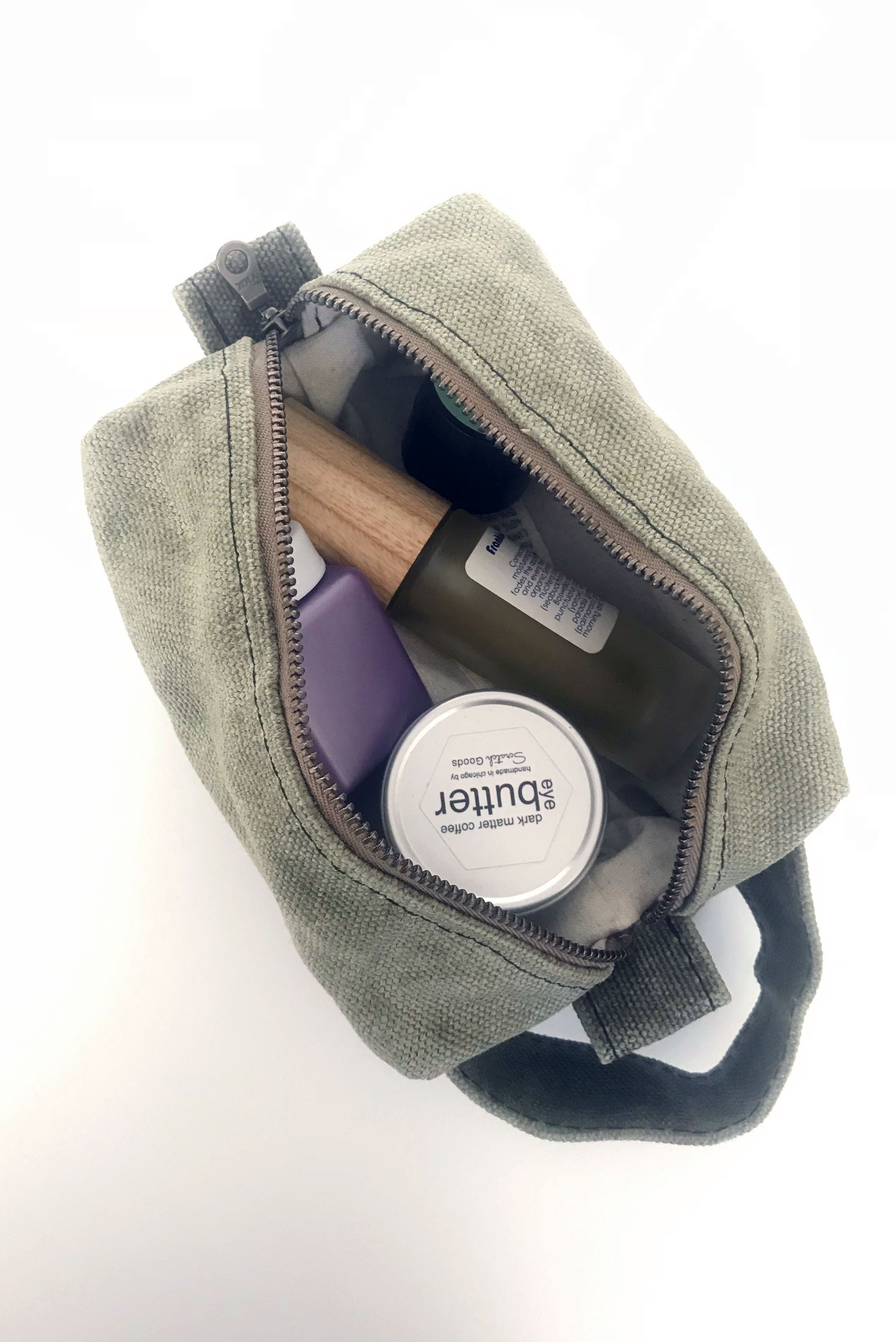 small waxed canvas toiletry bag with handle and brass zipper. The bag is open and contains small toiletries inside. The bag is sage green. 