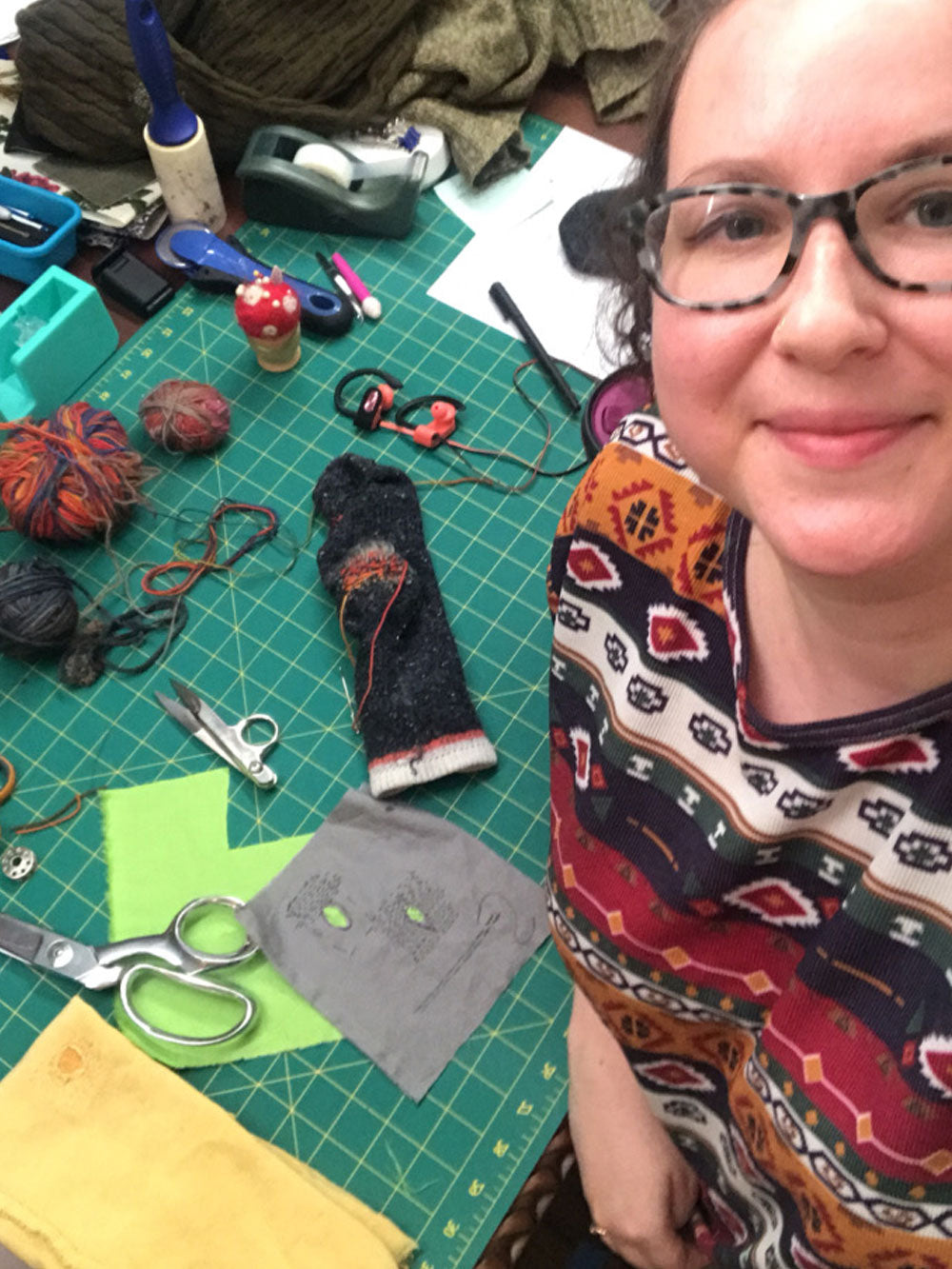 Woman smiles up at camera. Behind her a work table with thread, scissors, sewing accessories, and visible mending examples are on the table. 