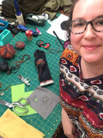 Load image into Gallery viewer, Woman smiles up at camera. Behind her a work table with thread, scissors, sewing accessories, and visible mending examples are on the table. 
