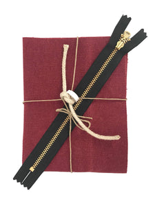 A small piece of folded red waxed canvas, a brass zipper, hemp rope, and role of leather tape, tied in a bow with twine. 