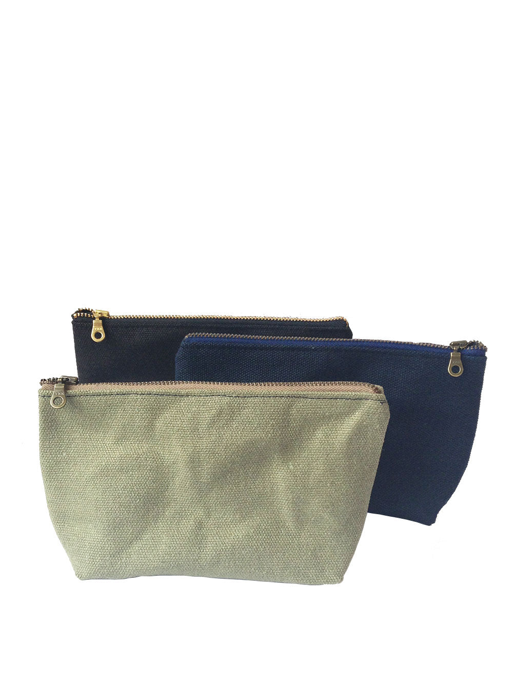 A row of three waxed canvas bags with brass zippers. Black in the black, then navy blue, then sage green on a white background. 